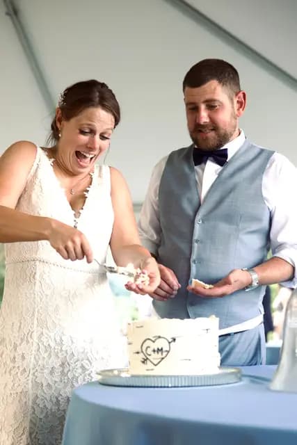 Bride laughs while cutting the wedding cake with the groom under the tent