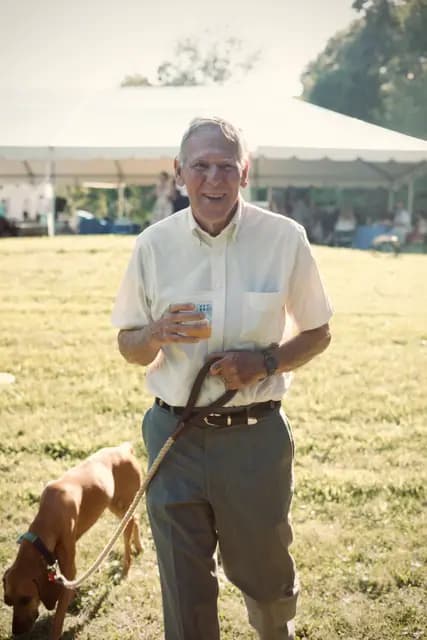 Smiling older guest holds a drink while walking a leashed dog by the tent