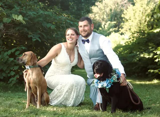 Smiling newlyweds kneel in a garden with their two festively dressed dogs