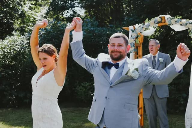 Newlyweds raise their joined hands in celebration beside the ceremony arch