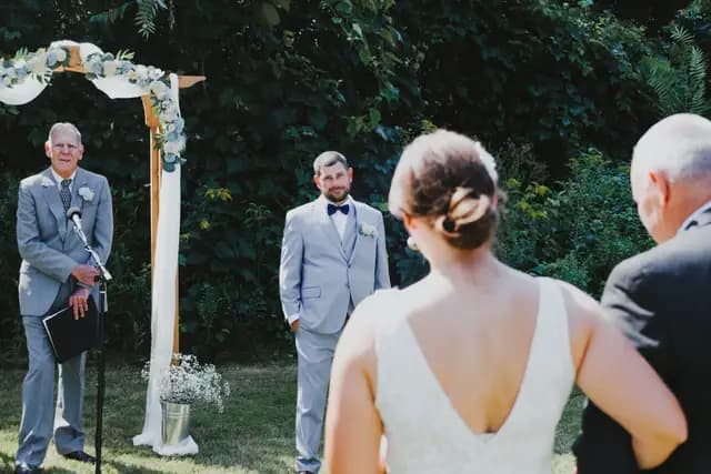 Officiant speaks at the arch as the groom watches the bride arrive