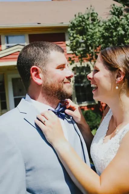 Bride laughs while adjusting the groom's bow tie outside a house