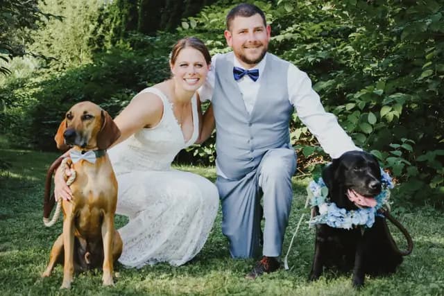 Bride and groom crouch in the grass with their two dogs dressed for the wedding
