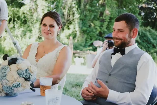 Bride and groom smile at their table as a guest photographs them