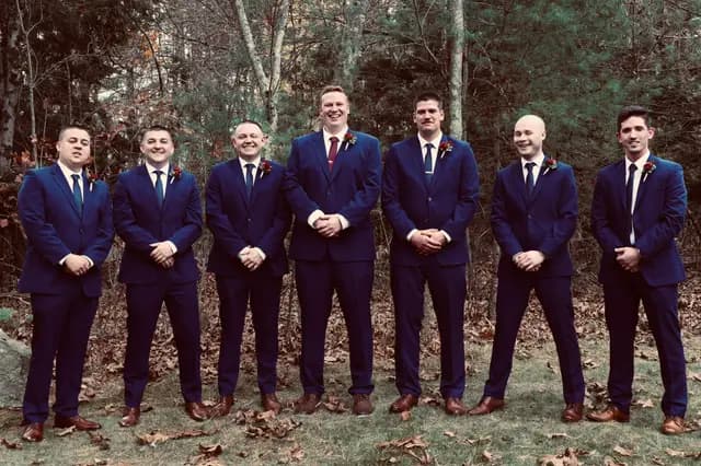 Seven groomsmen in navy suits smile together in the autumn woods