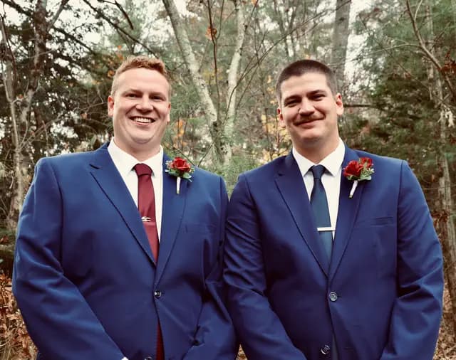 Groom and best man stand together with red rose boutonnieres