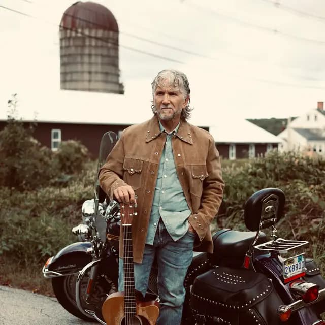 The artist leans on his guitar neck beside the studded saddlebags