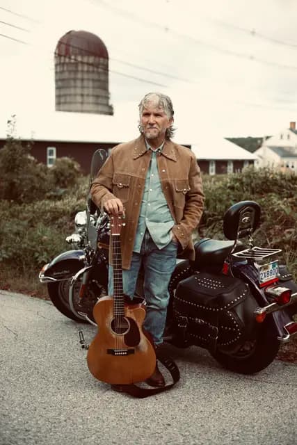 Musician in a suede jacket rests a guitar beside his Harley, farm silo behind