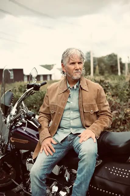 Seated on the Harley in a studded suede jacket, farmhouse behind