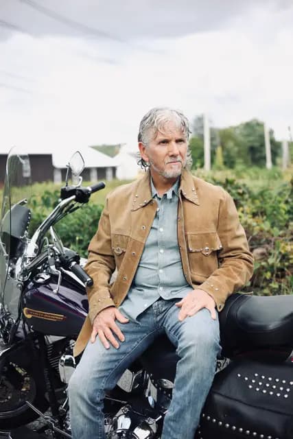 Hands on knees astride the Harley, white farmhouse in the haze
