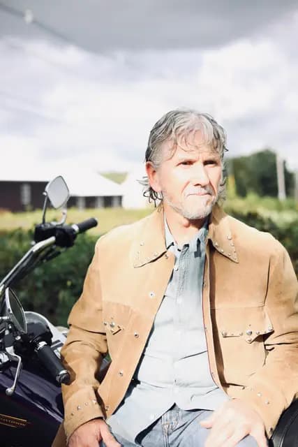 Seated portrait with a level stare, hedgerow behind