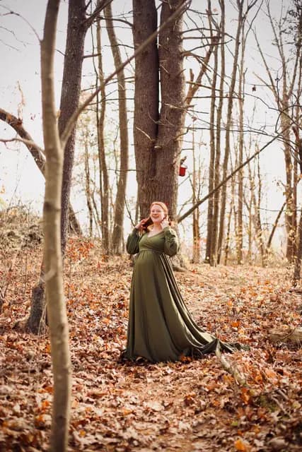 Expectant mother in a flowing olive gown laughs among bare autumn trees