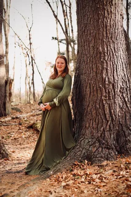 Leaning against a broad oak trunk with hands folded on her bump