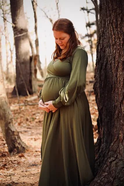 Looking down at her hands cradling the bump beside a rough trunk